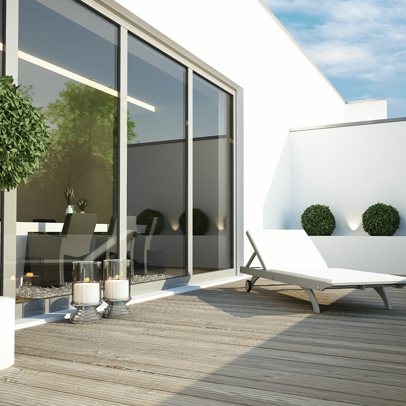 Terrasse extérieure design avec deck en bois, grandes portes vitrées, chaise longue blanche et arbustes décoratifs.