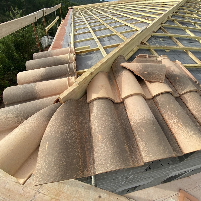 Toit en construction avec charpente bois, membrane respirante, tuiles canal posées et empilées, quelques tuiles cassées.